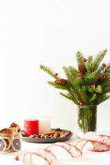Candles with dried slices of citrus fruits, hazelnuts with cinnamon, fir tree branches and cones in a vase on white wall background, new year and christmas concept