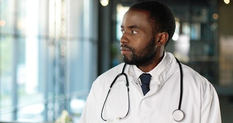 Fototapeta premium Portrait of African American young man doctor with stethoscope looking at camera and smiling cheerfully. Handsome male physician smile. Medic in white gown. Indoors.