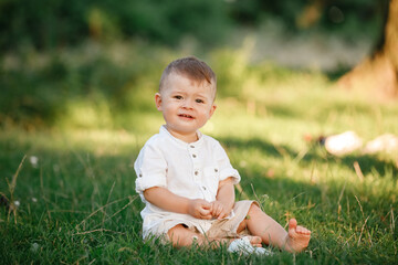 Happy adorable baby boy sitting on the grass in the park on summer day. Child in trendy and cute clothes