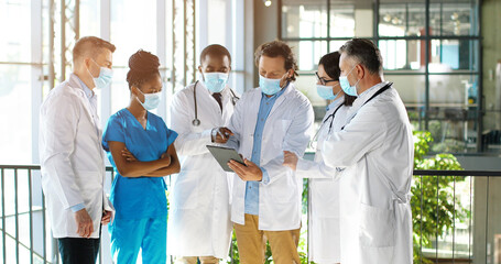 Male head of clinic standing with team of mixed-races doctors colleagues and using tablet device while watching on screen. Group of multi ethnic medics, men and women talking and having discussion.