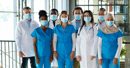 Mixed-races busy team of male and female doctors posing to camera and crossing hands in hospital. International group of medics in medical masks. Protected multi ethnic physicians and nurses in clinic