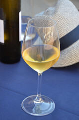 White wine in a crystal glass on a blue covered table, wine bottle and straw hat behind, outdoor in...