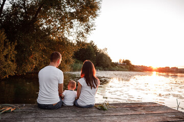 Young parents and their little son sitting on the wooden pier near the lake, at sunset on summer day.