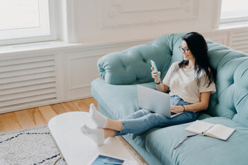 Horizontal shot of woman freelancer earns money online, checks email on smartphone, surrounded by modern technologies, organizer for making notes, uses high speed internet, sits on comfortable couch