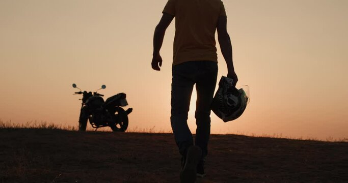 Motorcyclist with helmet in hand goes to his motorcycle at sunset. Slow motion shot