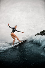 young woman confidently stands on the wake surf board and rides the wave.