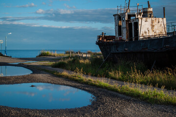 Fototapeta premium old rusty white ship, boat on grassy shore in light of sun, near round puddles on coast of lake Baikal, blue sky