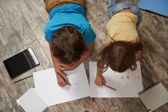 Siblings friendship. Top view of little caucasian brother and sister lying on the wooden floor at home and drawing together on white sheets of paper with colorful pencils