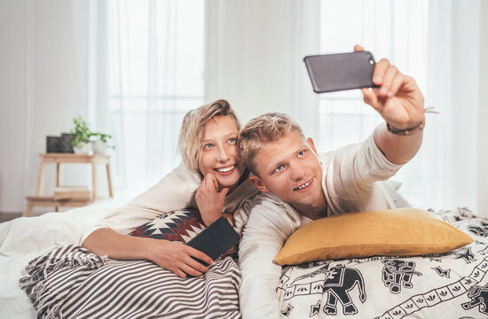 Cheerful young adults сouple in pajamas taking a selfie photo using a modern smartphone as they lazy relaxing lying in a cozy bed in the bedroom. . Couples relations concept image.