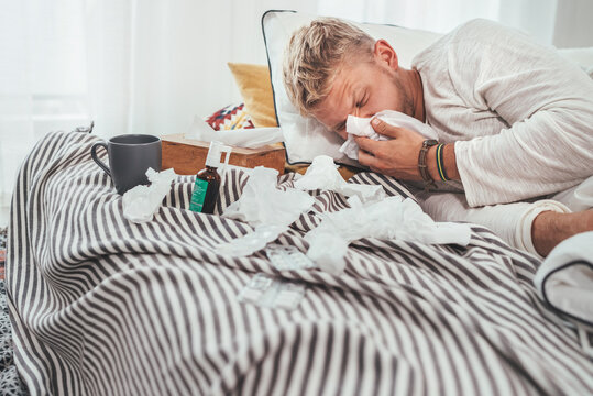 Unhealthy and tired man sneezing or wiping snotty nose man lying in a cozy home bed beside a lot of used paper tissues and medicines on the blanket. Season virus flu and home quarantine concept image.