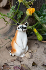 Racoon porcelain figurine on the ground with pumpkin plants and a wheel, autumn leaves