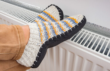 A woman is dressed in warm knitted woolen slippers  warm her legs on a heating radiator. The...