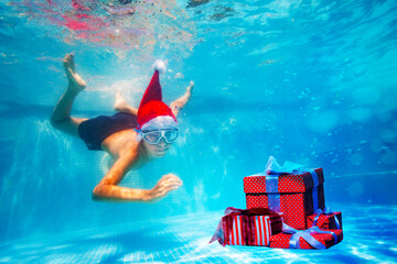 Happy boy in mask dive underwater in Santa Claus hat in the pool reaching for Christmas presents boxes on New Year vacation