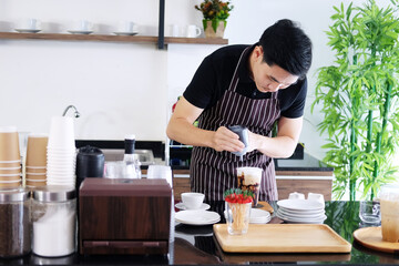 Asian barista young man pouring fresh Chocolate art in a plastic glass of Cappuccino iced coffee for according to order at counter bar in the morning at modern cafe and coffee shop.