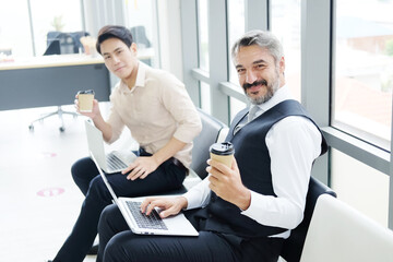 Smiling Caucasian Senior businessman drinking hot coffee with young Asain businessman and working with laptop and sitting distance together in workspace at office. New normal lifestyle concept.