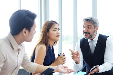 Smiling Asian team and Senior Caucasian boss is businesspeople teamwork relaxing and brainstorm. Businesswoman holding coffee glass and talking about smartphone at office. Focus at caucasian man.