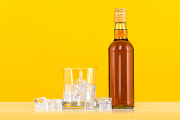 Glass of whiskey with ice cubes and bottle on yellow background