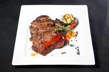 large grilled meat steak on a white square plate with a grill chapel and vegetables on a dark background
