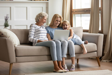 Funny movie. Happy family of 3 diverse age female members resting on sofa at home with laptop, hoary aged grandmother, her adult daughter and small grandchild girl watching comedy on computer screen