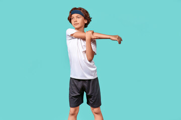 Stretching exercises. Teenage boy in sportswear looking away while warming up before workout or training, standing isolated over blue background