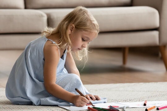 Talented artist. Inspired attentive little preschool child girl sitting on warm floor in cozy apartment painting funny pictures with fiber-tip pens, drawing bright imaginary world on piece of paper