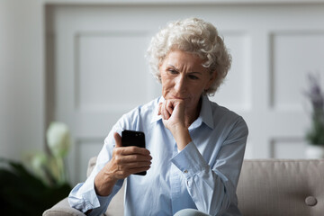 So what should I press next. Thoughtful old lady sits on sofa holding cellphone and looking pensive...