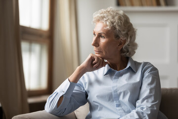 Lost in thoughts. Unhappy elderly female sitting on couch at home alone propping chin with hand and looking aside with pain worrying for children, troubled by hard present day life or health problems