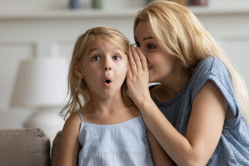 It is a great unknown. Smiling adult mom telling cute story as big mystery on ear of surprised little daughter, creative young nanny finding way to heart of small preschool child sharing funny secret