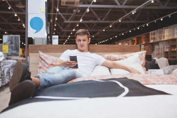 Attractive male customer trying new bed at furniture store, lying on it using his phone