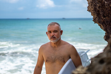 Portrait of handsome shirtless man surfer, holding white surf board  and cliff, rocks, blue wave on background