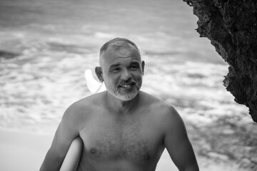 Black and white portrait of handsome shirtless man surfer , holding white surf board  and cliff, rocks on background