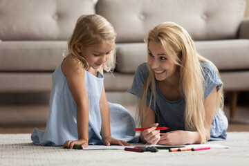 Fototapeta premium Creative activity. Happy smiling young mother spending leisure time with little kid daughter lying on heated floor in living room drawing funny pictures with bright colorful marker pens in paper album