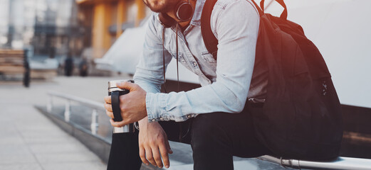 Modern smiling bearded hipster with black backpack, headphones on neck and thermo mug with coffee...