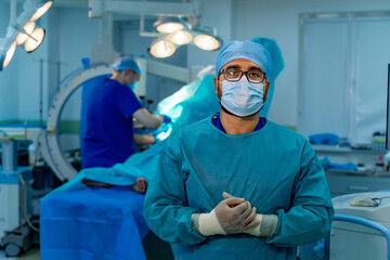 Medic in blue scrubs wearing face mask, glasses and latex gloves. Operating theatre. Modern surgery equipment.