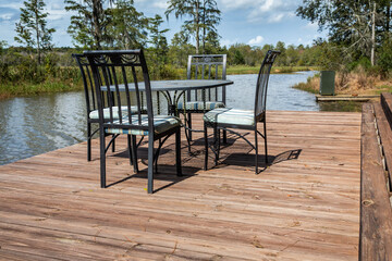 A set of black metal chairs and a table sitting on a large wooden dock deck outside of a hunting and fishing cabin near a large pond in Georgia for outdoor dining
