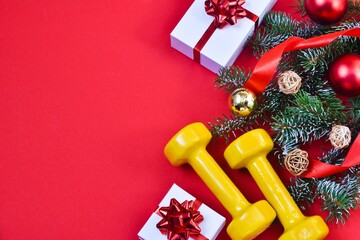 Christmas sports equipment. Dumbbells, red ribbon, fir branch and white gift box with a red bow on a red background. Healthy lifestyle and sport concept. View from above. Copy space