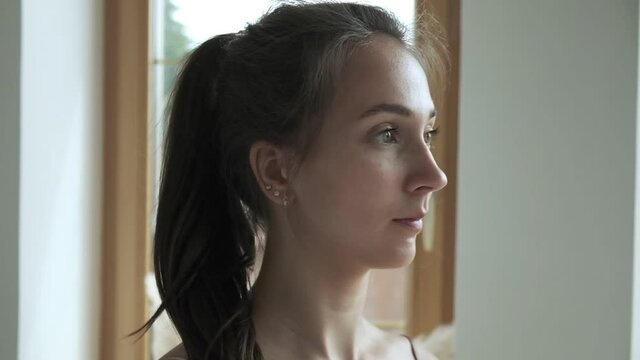 sporty brunette pretty female stands at window, fixes her hair, collects hair in bun. closeup shot of young caucasian woman standing in living room