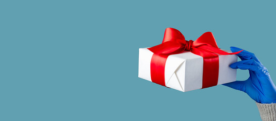 Christmas covid-19. Gift surprise. Safety delivery. Female hand in medicine gloves holding present box taped red ribbon isolated on blue copy space. New Year 2021. Winter holiday.