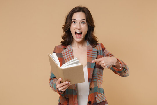 Shocked surprised young brunette woman 20s wearing casual checkered jacket holding reading pointing index finger on book looking camera isolated on pastel beige colour background, studio portrait.