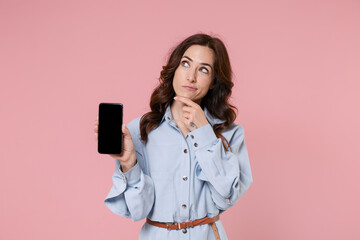 Pensive young brunette woman 20s in casual blue shirt dress hold mobile phone with blank empty screen mock up copy space put hand prop up on chin isolated on pink colour background, studio portrait.