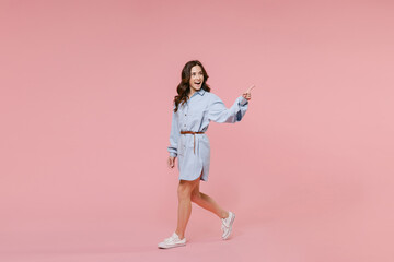 Full length side view portrait of excited young brunette woman 20s wearing casual blue shirt dress pointing index finger aside on mock up copy space isolated on pastel pink colour background studio.