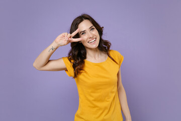 Fototapeta na wymiar Smiling cheerful funny beautiful young brunette woman 20s wearing basic yellow t-shirt posing standing showing victory sign looking camera isolated on pastel violet colour background, studio portrait.