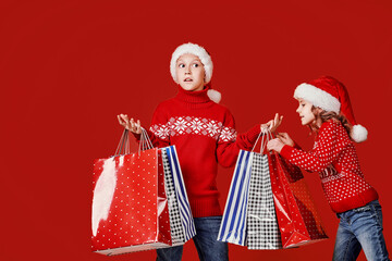 Cute little girl in Santa hat hanging shopping bag with Christmas gifts of astonished boy during...