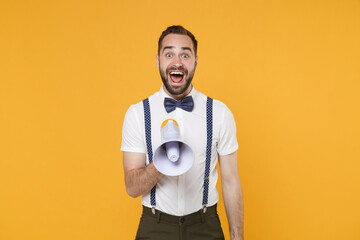 Surprised excited young bearded man 20s wearing white shirt bow-tie suspender posing standing screaming in megaphone looking camera isolated on bright yellow color wall background studio portrait.