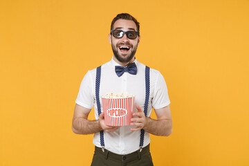 Surprised young bearded man 20s wearing white shirt bow-tie suspender 3d glasses posing watching movie film, holding bucket of popcorn isolated on bright yellow color wall background studio portrait.