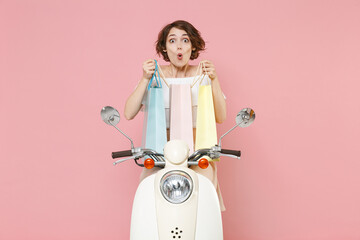 Shocked amazed young brunette woman 20s wearing white summer clothes hold package bag with purchases after shopping sitting driving moped isolated on pastel pink colour background, studio portrait.