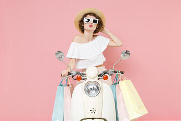 Pretty young woman in white summer clothes hat glasses hold package bag with purchases after shopping blowing sending air kiss driving moped isolated on pastel pink colour background studio portrait.