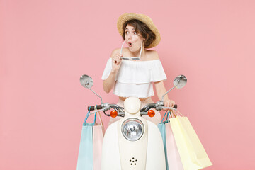 Shocked young woman 20s wearing white summer clothes hat glasses hold package bag with purchases after shopping looking aside driving moped isolated on pastel pink colour background studio portrait.