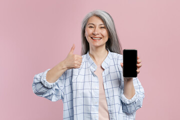 Smiling gray-haired asian woman in white checkered shirt showing thumb up hold mobile cell phone with blank empty screen mock up copy space isolated on pastel pink colour background, studio portrait.