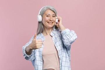 Smiling gray-haired asian woman wearing basic white checkered shirt standing listening music with headphones showing thumb up looking camera isolated on pastel pink colour background, studio portrait.